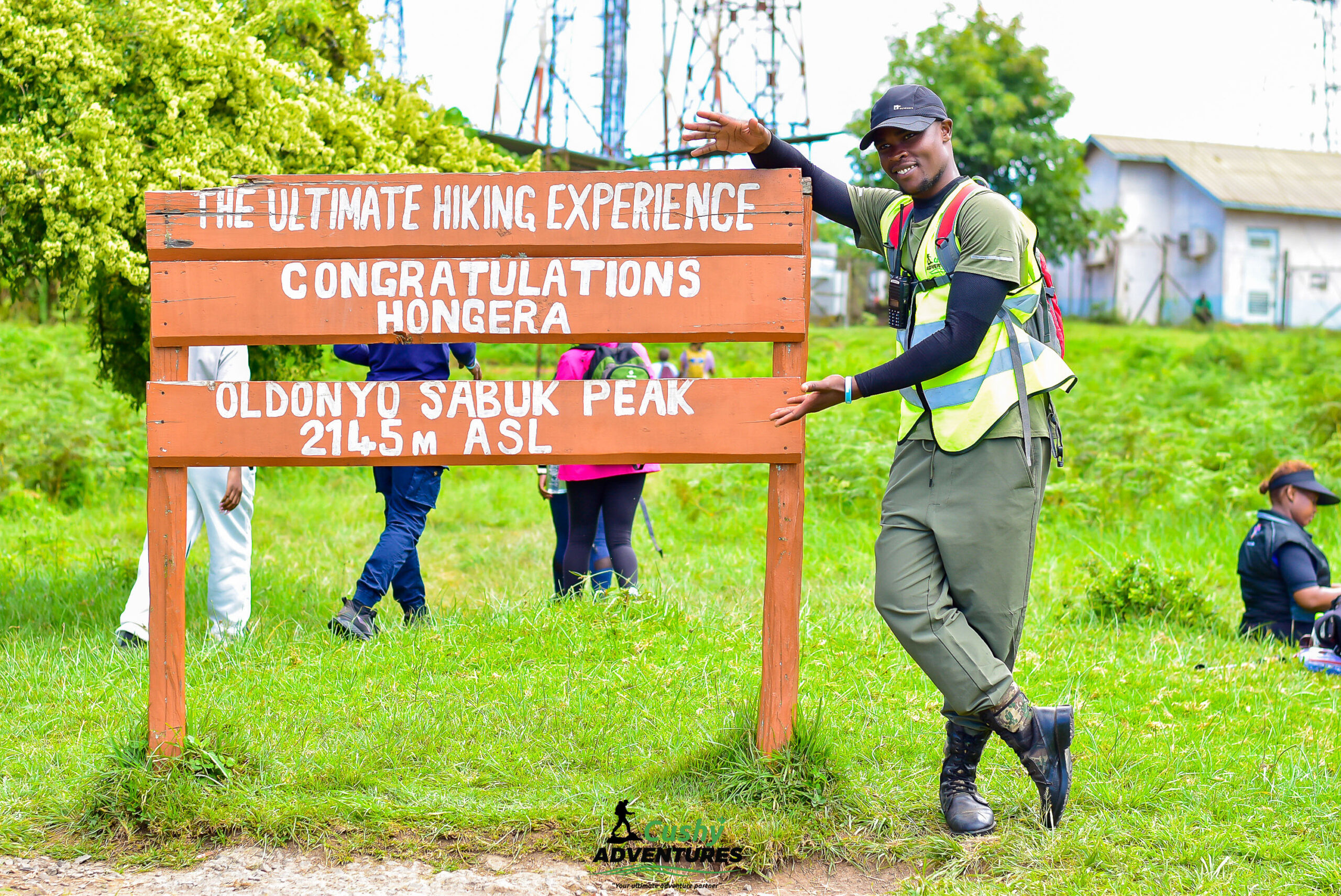 Company hiking guide standing at Mount Kilimambogo summit board, smiling after a successful hike.