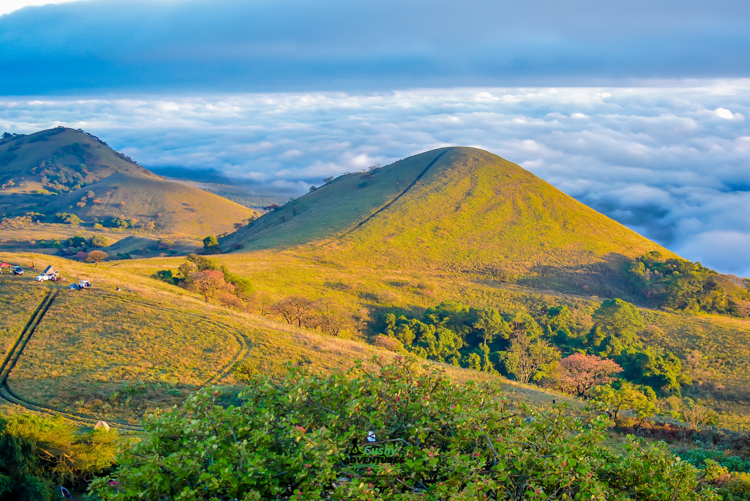 Green rolling hills of Chyulu Hills rising above a sea of clouds at sunrise, with tents and vehicles visible in the distance.